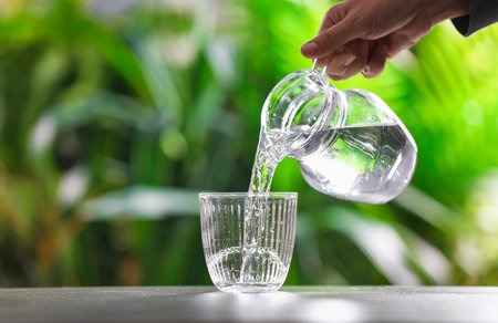 Woman pouring water into glass from jug at gray table against blurred green background, closeup....