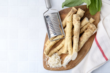 Grated horseradish, grater, leaves and fresh roots on white tiled table, flat lay. Space for textの写真素材