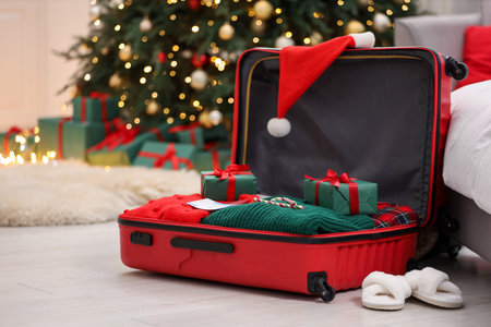 Christmas vacation. Suitcase with Santa hat, clothes, candy canes and gift boxes on floor indoors, closeupの写真素材