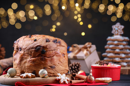 Christmas food. Delicious Panettone cake, gingerbread cookies, candied fruits and decor on black wooden table against blurred lights, closeup. Bokeh effectの写真素材