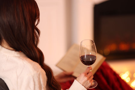 Woman with red wine reading book near fireplace at home, closeupの写真素材
