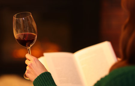 Woman with red wine reading book near fireplace at home, closeupの写真素材