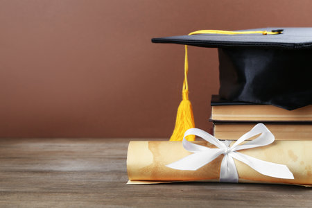 Graduation cap, diploma and books on wooden table against brown background, closeup. Space for textの写真素材