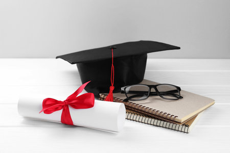 Graduation cap, diploma, notebooks and glasses on white wooden table against light backgroundの写真素材