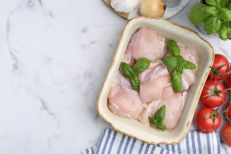Raw chicken thighs with basil in baking dish and products on white marble table, flat lay. Space for textの写真素材
