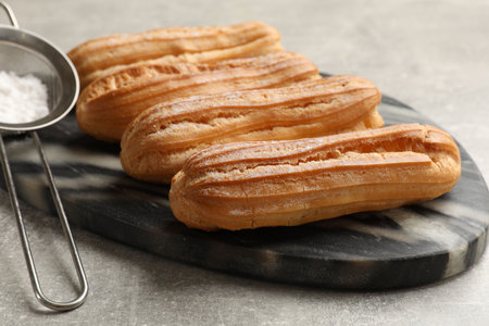 Delicious eclairs and strainer with powdered sugar on light gray table, closeup