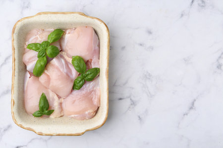 Raw chicken thighs with basil in baking dish on white marble table, top view. Space for textの写真素材