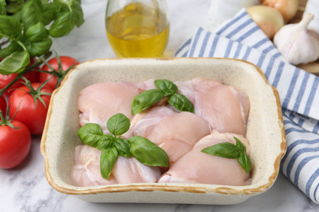 Raw chicken thighs with basil in baking dish and products on white marble table, closeupの写真素材