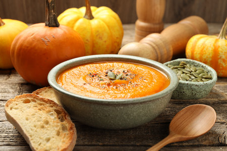 Tasty pumpkin cream soup with seeds in bowl, roasted bread, vegetables and spoon on wooden table, closeupの写真素材