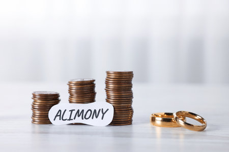 Paper note with word Alimony, coins and wedding rings on wooden table, closeupの写真素材