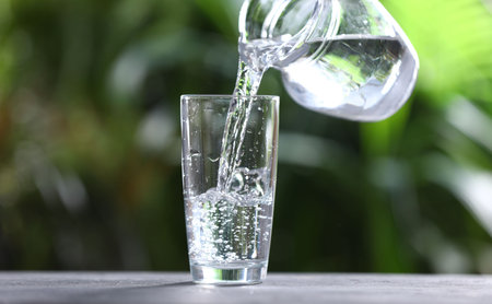 Pouring water into glass from jug at gray table against blurred green background, closeup