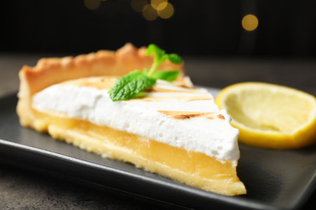 Slice of tasty lemon meringue tart with mint served on black table against blurred lights, closeupの写真素材