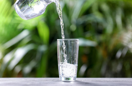 Pouring water into glass from jug at gray table against blurred green background, closeup