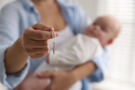 Sacrament of baptism. Woman holding little baby and gold cross indoors, selective focus