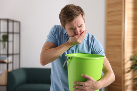 Young man with bucket suffering from nausea indoorsの写真素材