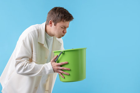 Young man with bucket suffering from nausea on light blue background, space for textの写真素材