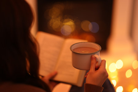 Woman with cocoa drink reading book near fireplace at home, closeup. Bokeh effectの写真素材