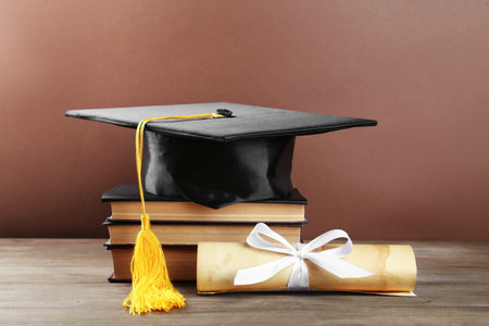 Graduation cap, diploma and books on wooden table against brown background, closeupの写真素材
