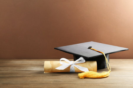 Graduation cap and diploma on wooden table against brown background. Space for textの写真素材