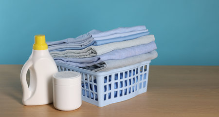 Laundry basket with clean clothes and detergents on wooden table against light blue background, space for textの写真素材