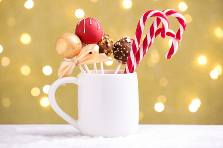 Tasty cake pops and candy canes in mug on white table against beige background with blurred lights. Christmas treatの写真素材