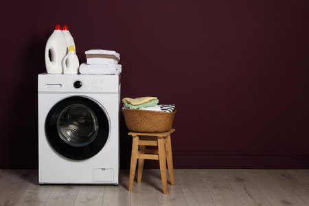 Modern washing machine, detergents and clothes near burgundy wall indoors, space for textの写真素材