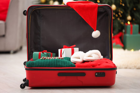 Christmas vacation. Suitcase with Santa hat, clothes and gift boxes on floor indoors, closeupの写真素材