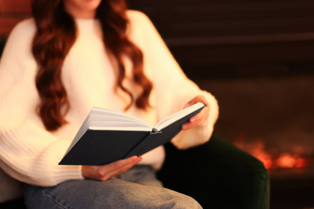 Woman reading book near fireplace at home, closeup. Space for textの写真素材