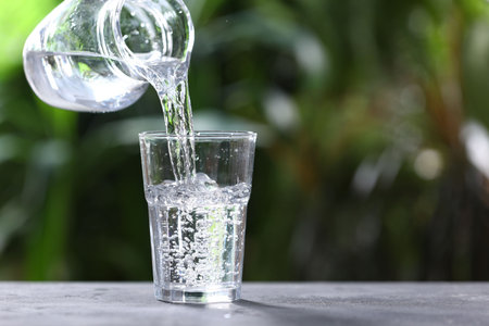 Pouring water into glass from jug at gray table against blurred green background, closeup. Space for textの写真素材