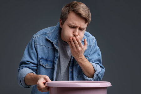 Young man with basin suffering from nausea on gray background