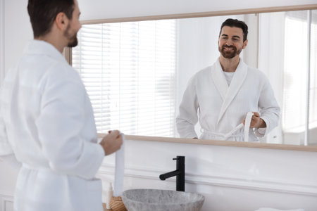 Handsome man wearing bathrobe and looking at himself in mirror indoorsの写真素材
