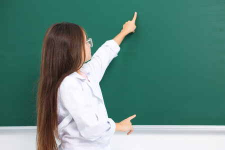 Little girl in laboratory coat and glasses pointing at something at green chalkboard indoors, space for text. Child and scienceの写真素材