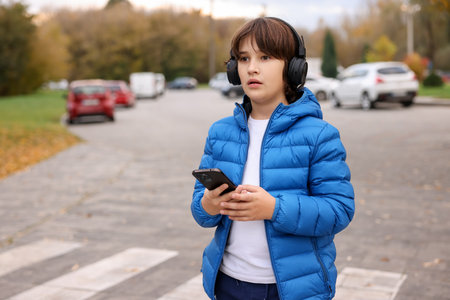 Little boy in headphones using smartphone on pedestrian crossing outdoors, space for text. Child in danger and road safetyの写真素材