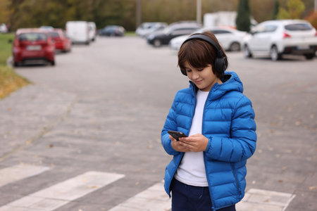 Little boy in headphones using smartphone on pedestrian crossing outdoors, space for text. Child in danger and road safetyの写真素材
