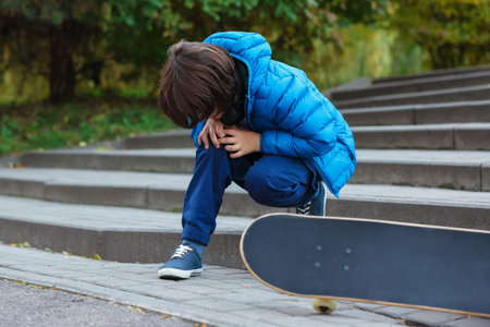 Little boy suffering from knee pain on stairs after falling from skateboard outdoors. Child in dangerの写真素材