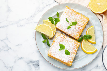 Tasty lemon bars with powdered sugar, mint and fresh fruits on white marble table, flat lay. Space for textの写真素材