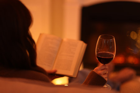 Woman with red wine reading book in armchair near fireplace at home, closeupの写真素材
