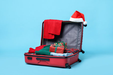 Christmas vacation. Open suitcase with Santa hat, clothes and gift boxes on light blue backgroundの写真素材