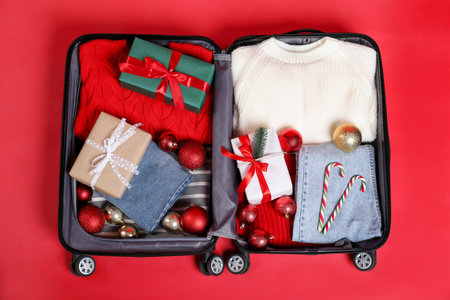 Christmas vacation. Open suitcase with clothes, gift boxes, baubles and candy canes on red background, top viewの写真素材