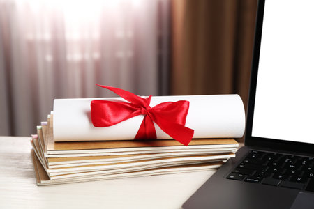 Diploma with red bow, stacked copybooks and laptop on white wooden table indoors, closeupの写真素材