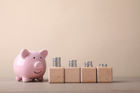 Empty cubes with stacked coins and piggy bank on wooden table against beige background. Space for designの写真素材