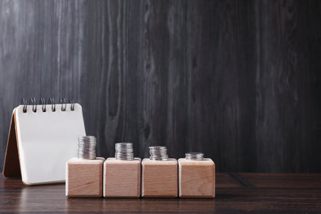 Empty cubes with stacked coins and notebook on wooden table against dark gray background. Space for...