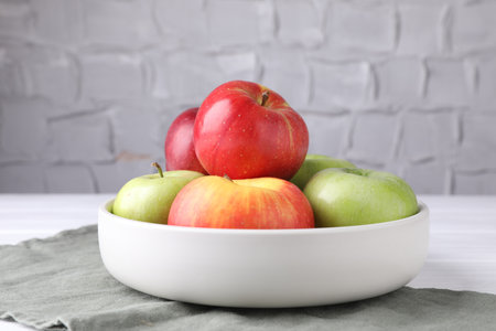 Fresh red and green apples in bowl on white table near gray textured wall, closeupの写真素材