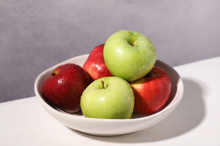 Fresh red and green apples with water drops in bowl on white table against gray background, closeupの写真素材