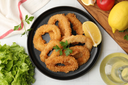 Deep fried squid rings with parsley and lemon slice on white tiled table, flat lay