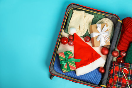 Christmas vacation. Open suitcase with Santa hat, clothes, gift boxes, baubles and candy canes on light blue background, top view. Space for textの写真素材