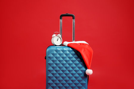 Santa hat and alarm clock on blue suitcase against red background. Christmas vacationの写真素材
