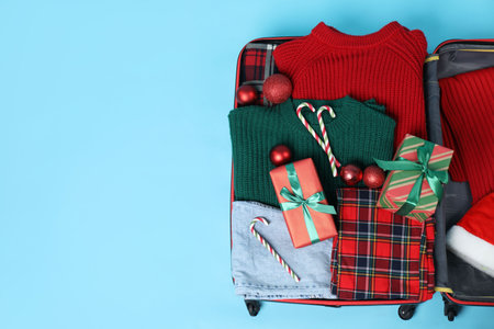 Christmas vacation. Open suitcase with clothes, gift boxes, baubles and candy canes on light blue background, top view. Space for textの写真素材