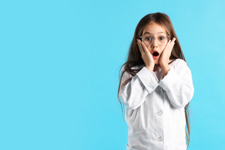 Surprised little girl in laboratory coat and glasses on light blue background, space for text. Child...