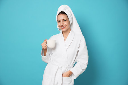 Woman in bathrobe with cup of coffee on light blue background